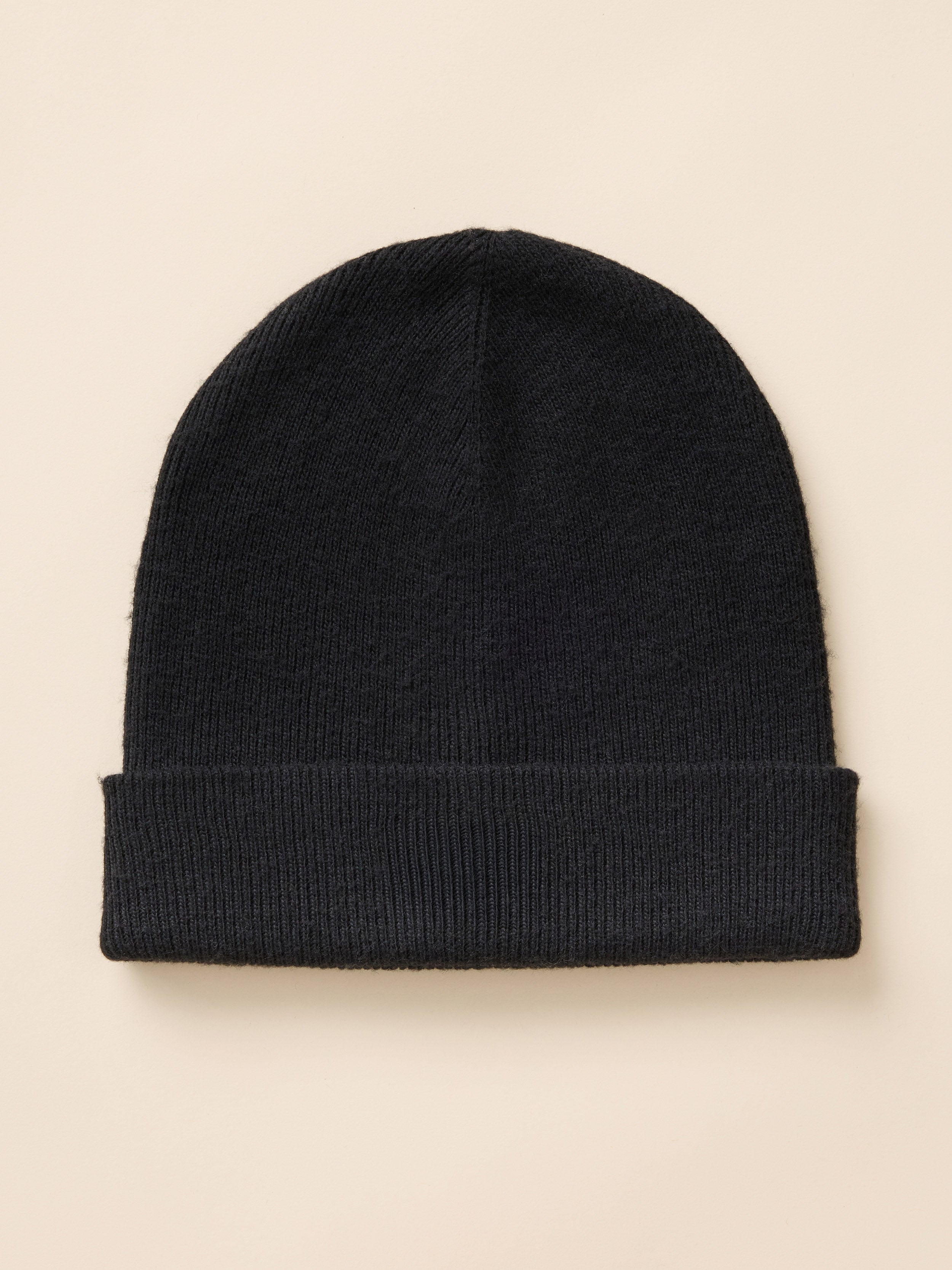 A black ribbed knit beanie hat with a folded cuff, photographed flat against a light beige background.