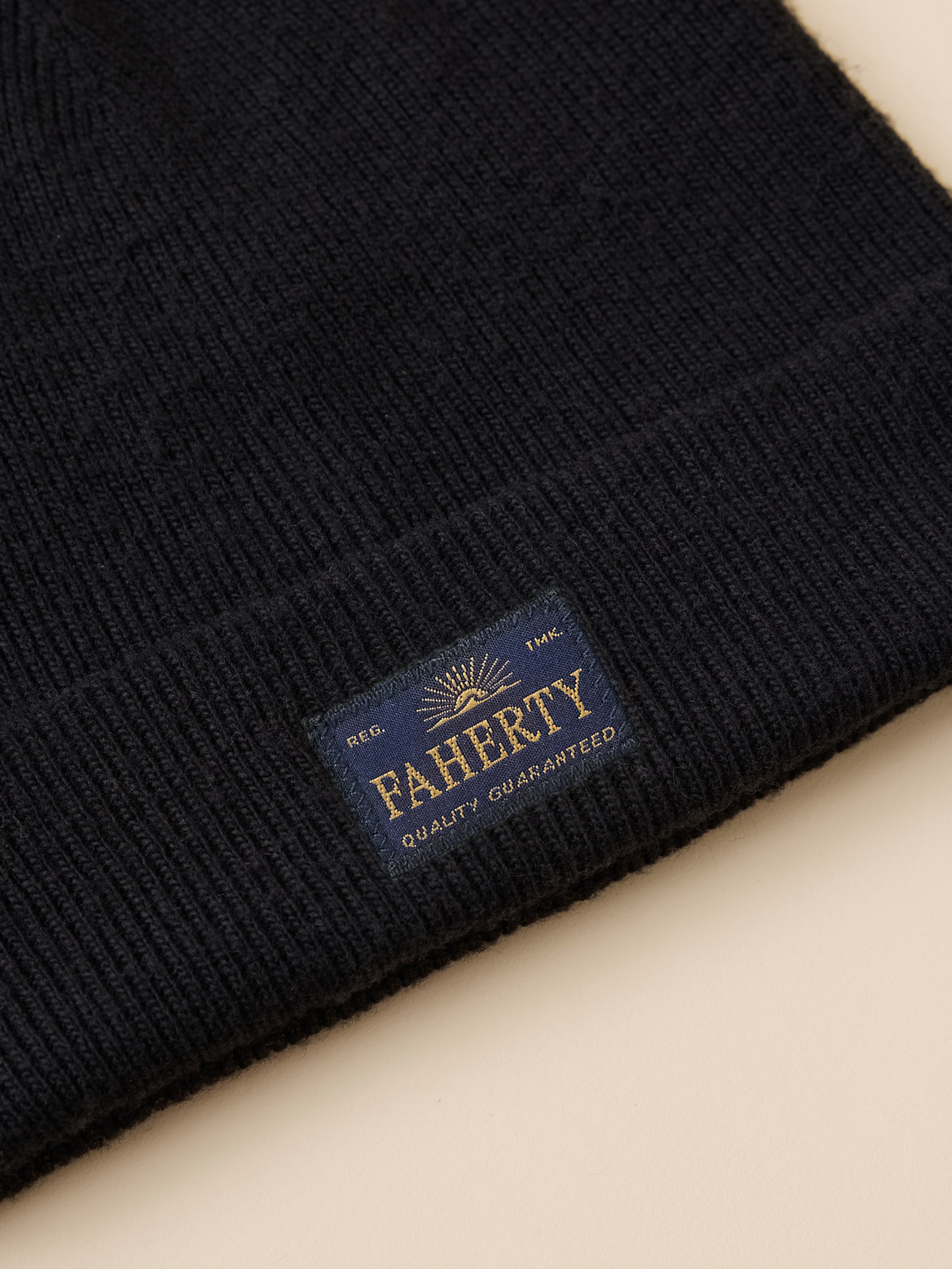 Close-up of a black ribbed knit beanie with a folded cuff showing a navy blue woven label that reads FAHERTY Quality Guaranteed.