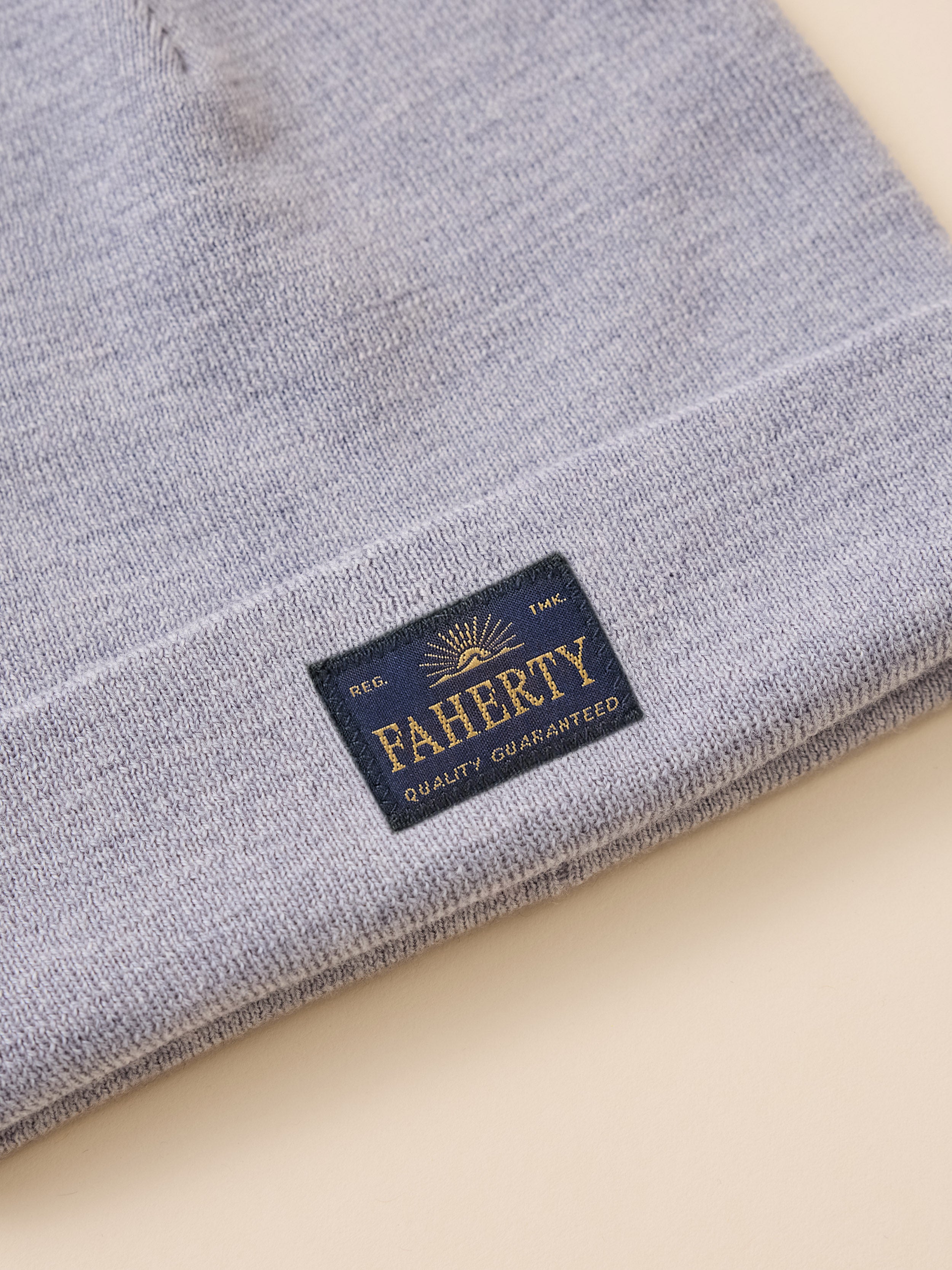 A close-up of a folded light heather gray knit item, likely a beanie or hat, showing a navy blue sewn-in label with gold lettering that reads FAHERTY and Quality Guaranteed.