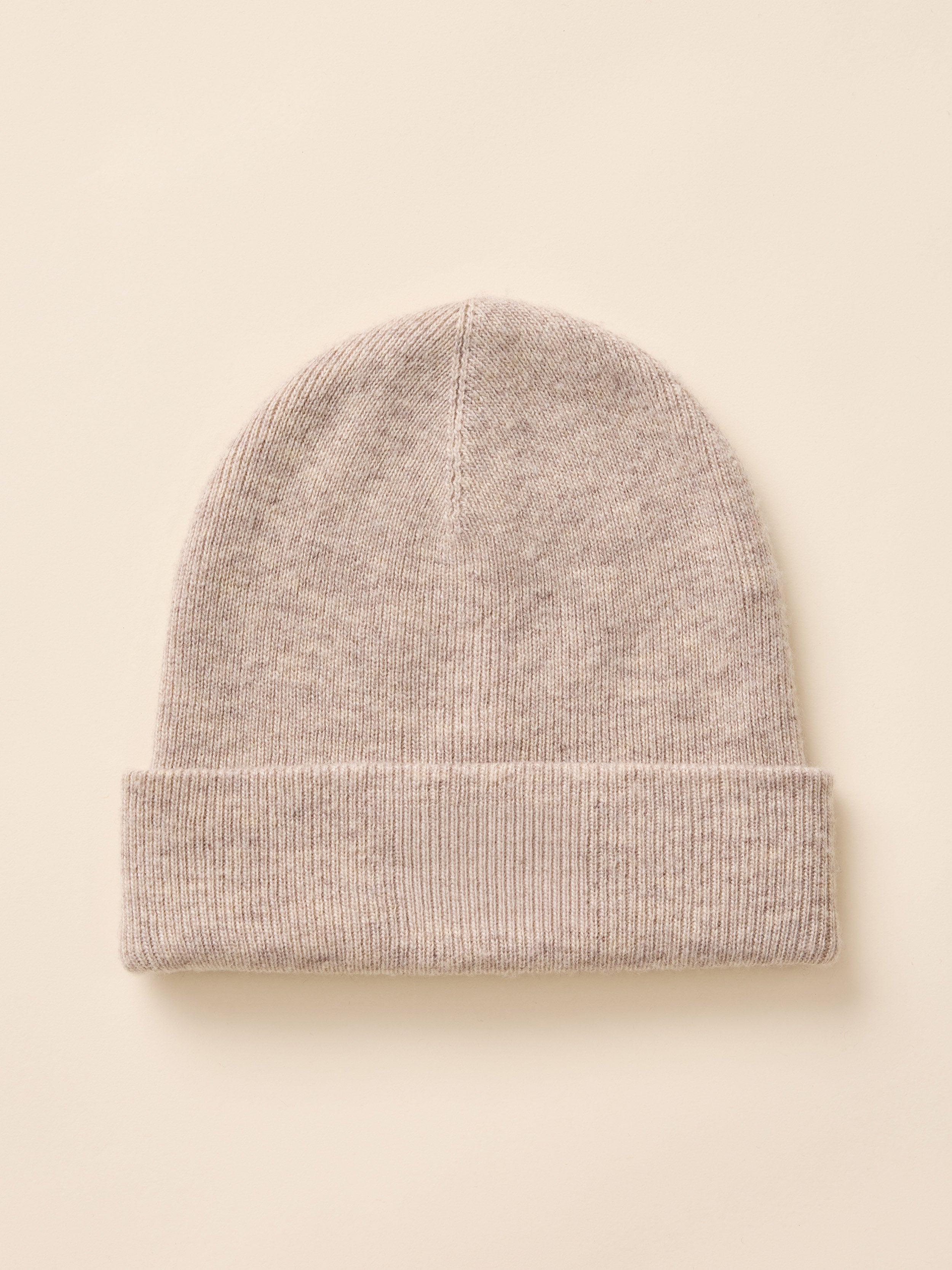 A light beige knit beanie hat with a folded cuff, shown flat against a solid cream background.
