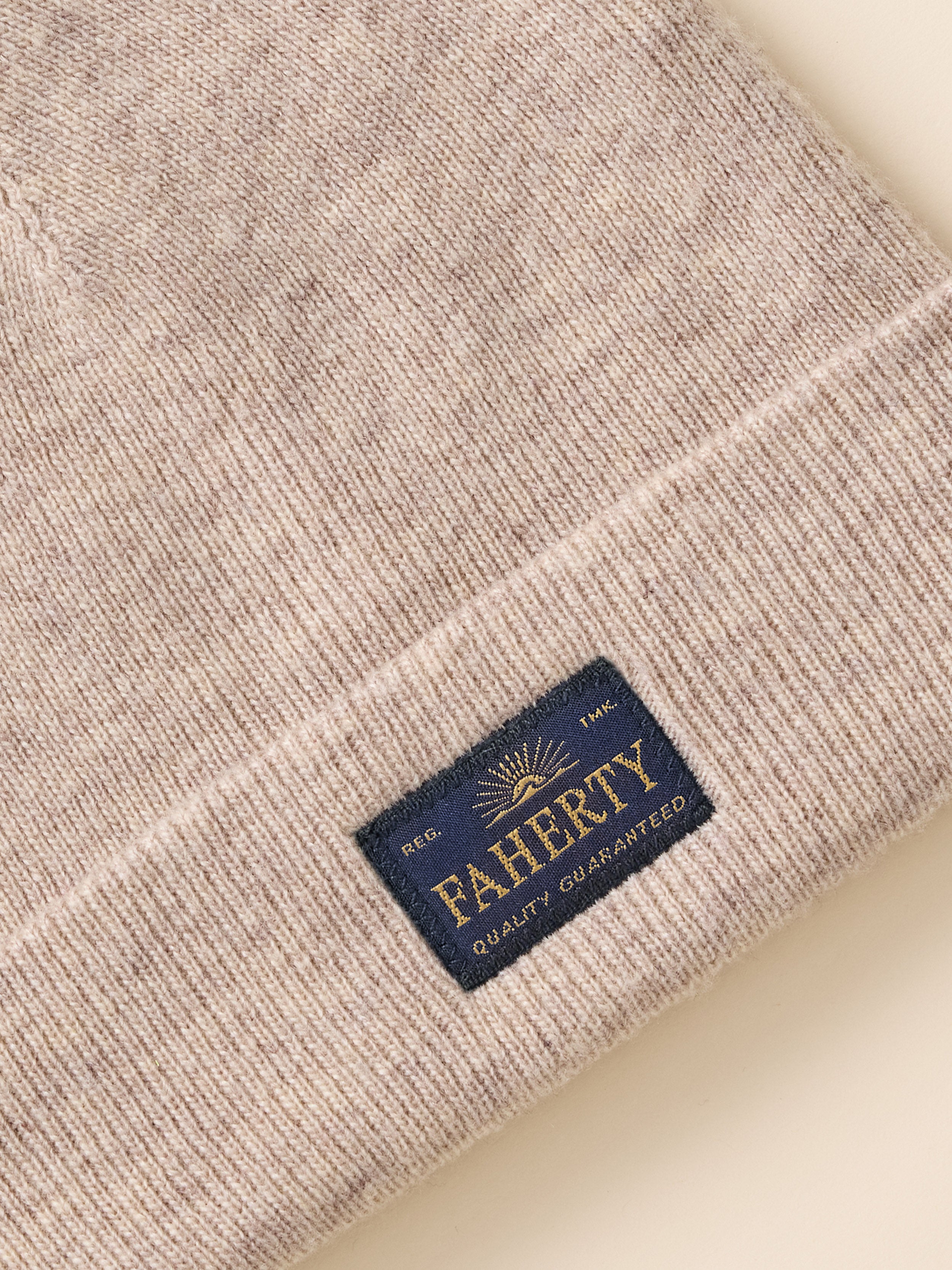 Close-up of a ribbed knit beanie in a heathered light beige or oatmeal tone, featuring a navy blue rectangular brand patch with gold lettering that reads FAHERTY and smaller text 'SEC. 1861.