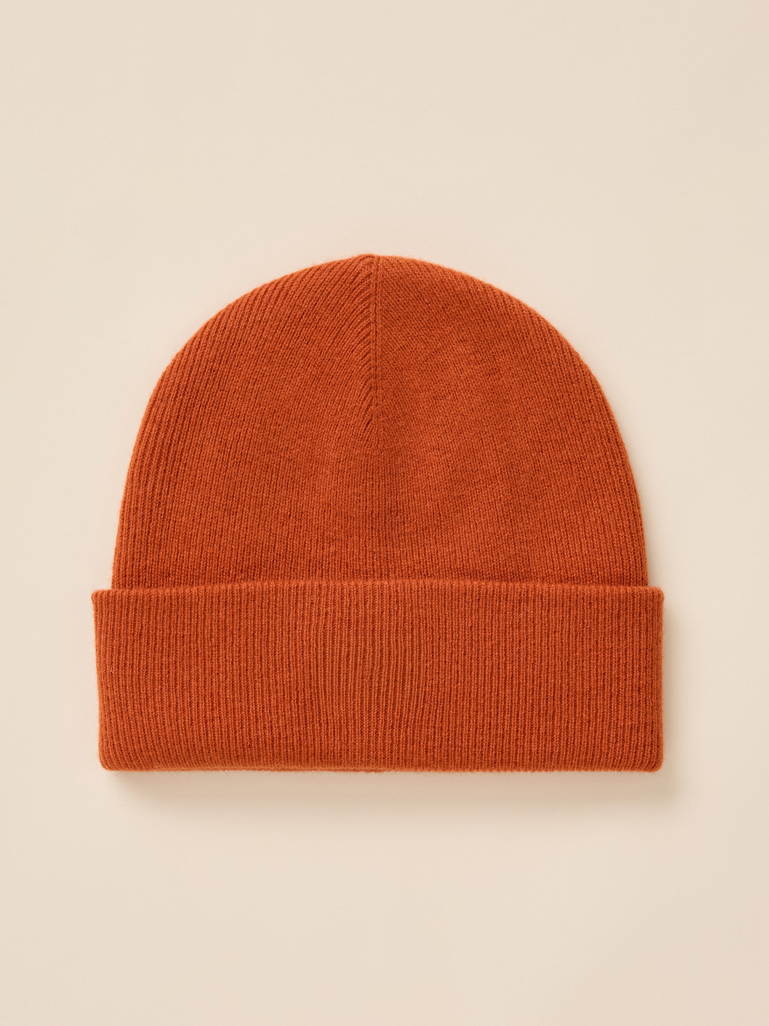 A classic ribbed knit beanie hat in a warm rust orange color, presented flat against a light cream background.