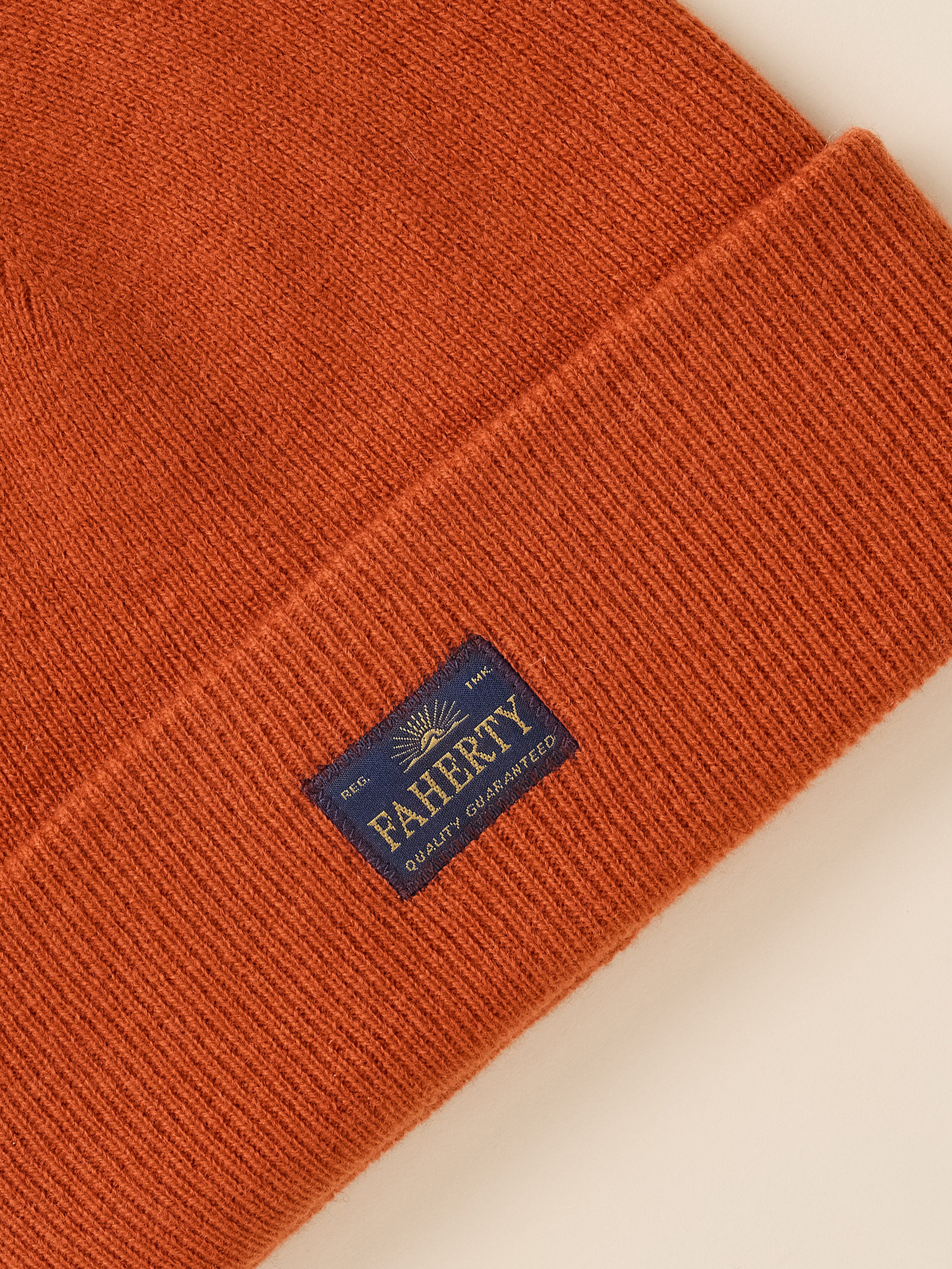 Close-up of a ribbed knit beanie in a warm rust orange color, featuring a navy blue brand patch that reads 'FAHERTY Quality Guaranteed' with a gold sunburst logo.
