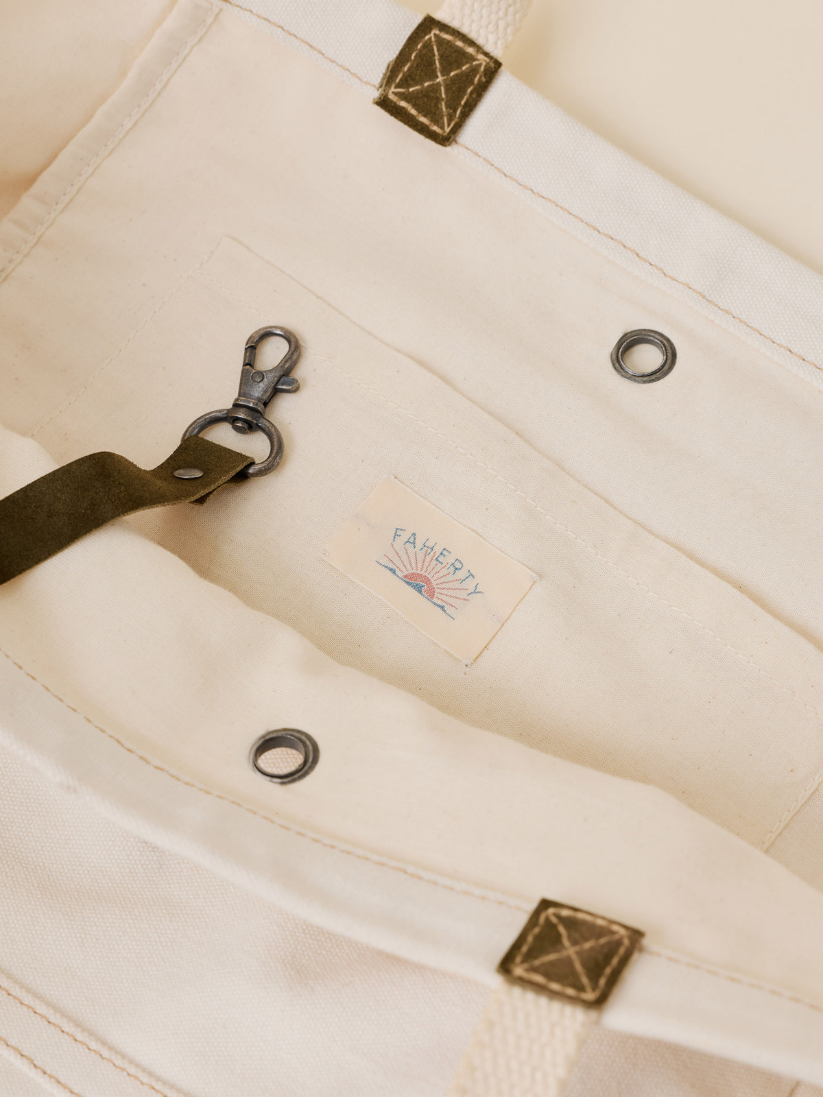 Close-up of a cream canvas tote bag showing a front pocket with metal grommets, a dark olive strap with a brass clasp, and a small sewn-in Faherty brand label.