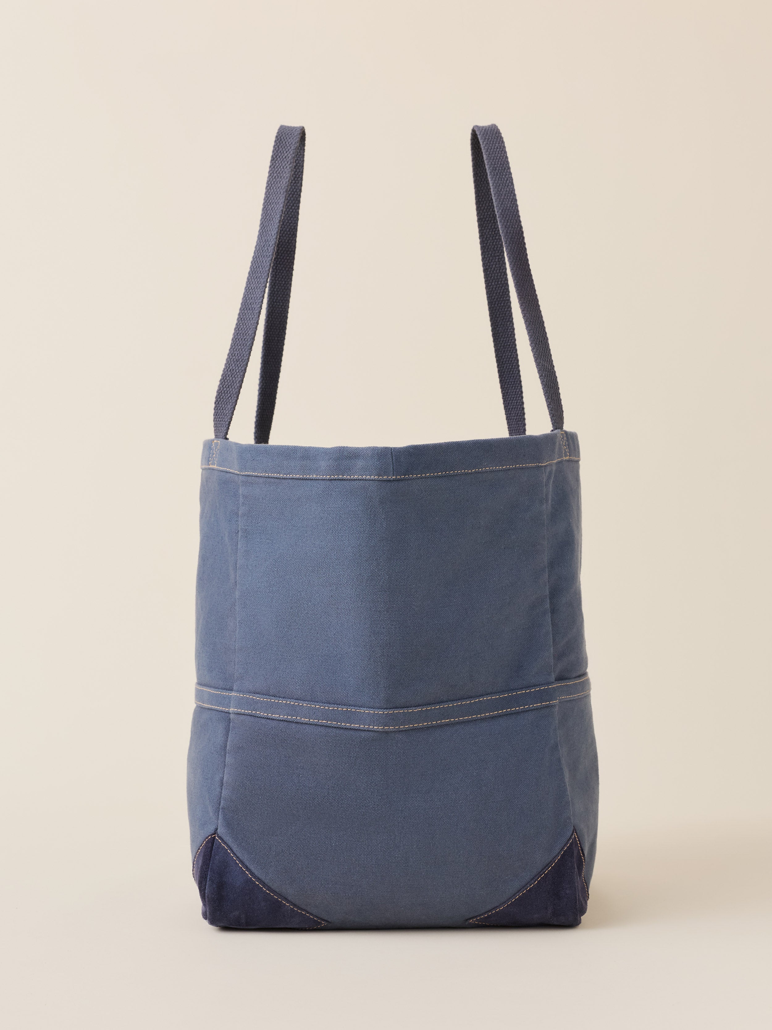 A blue canvas tote bag with dark navy blue bottom corners and shoulder straps, shown against a solid beige background.