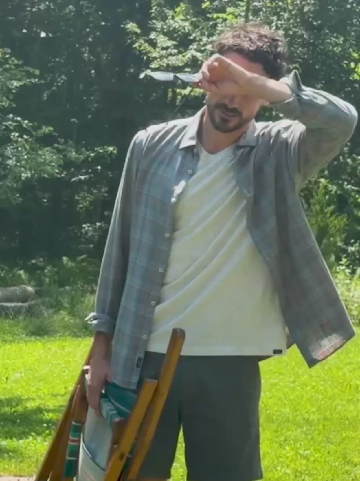 Man standing outside in the grass holding a chair while wearing a plaid shirt over a white tee and shorts