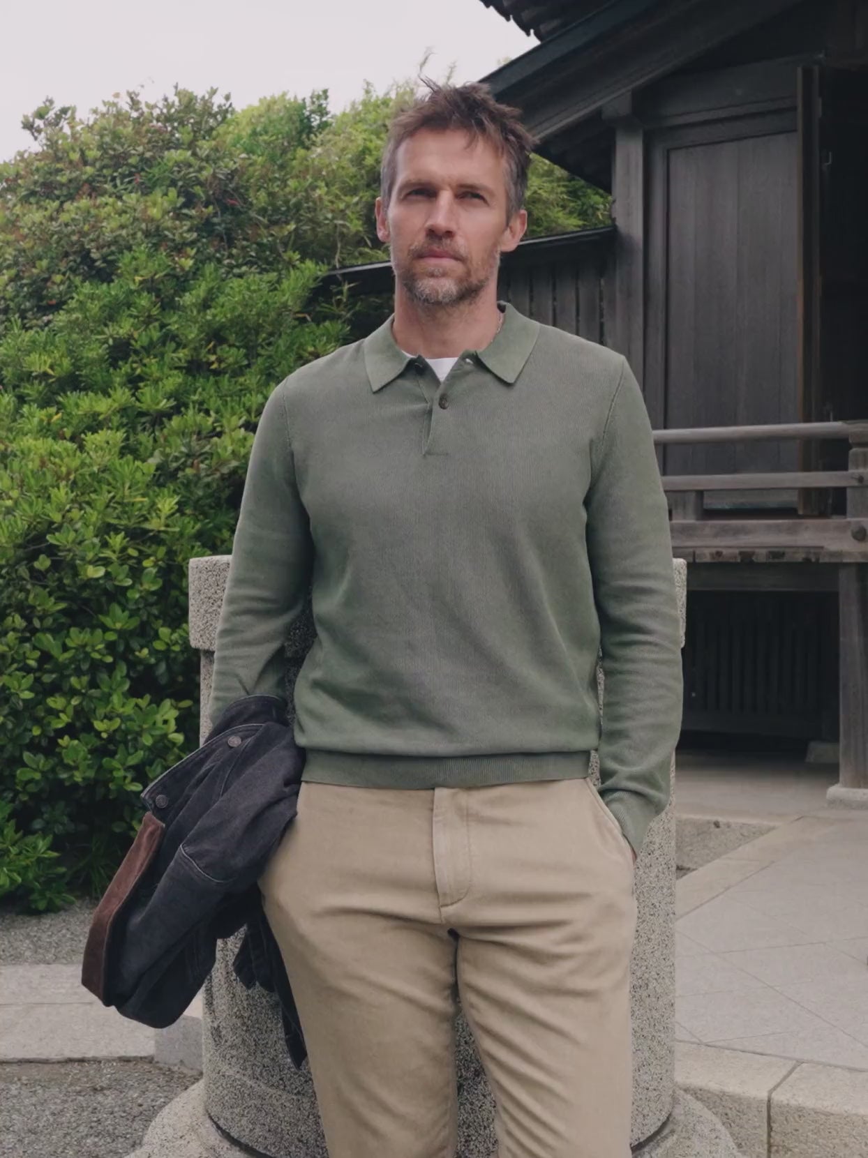 Video of a man wearing a green long sleeve sweater polo and chino pants standing outside a japanese house holding a jacket