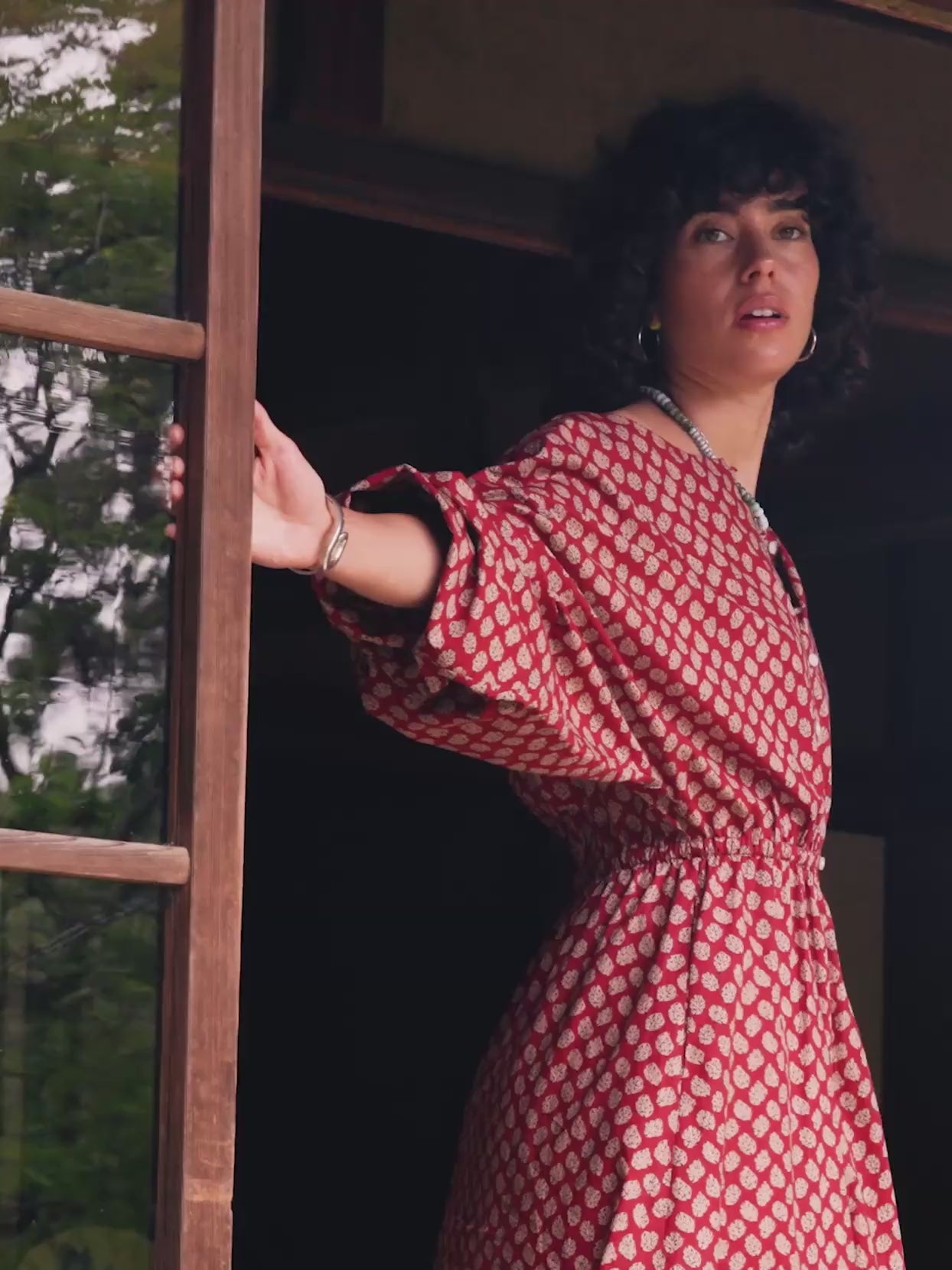 Video of a woman with curly dark hair models a rust-red off-the-shoulder maxi dress with a white floral pattern, set in a traditional Japanese room with shoji screens.
