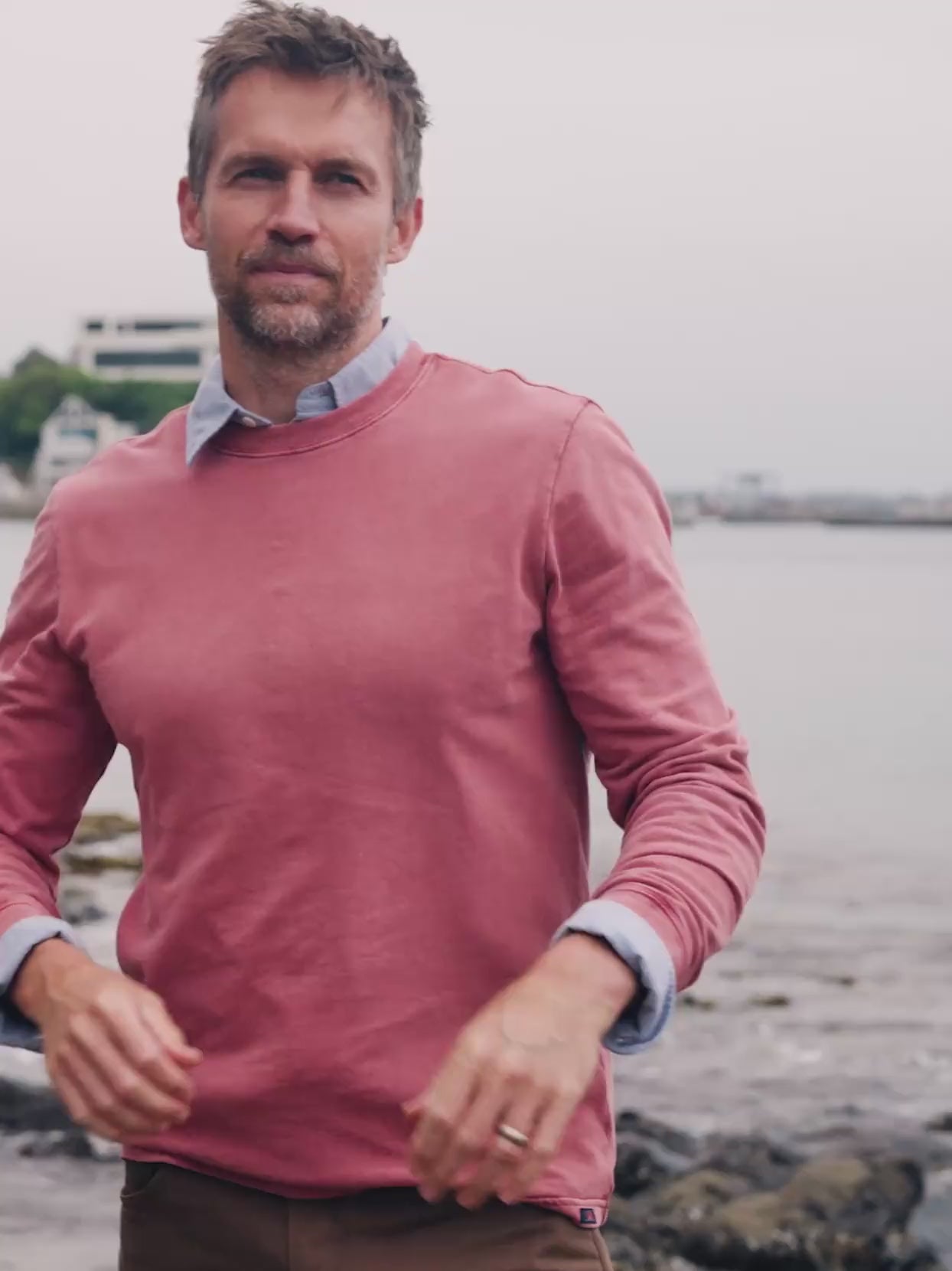 Video of a man wearing a red shirt over a blue button down shirt standing near the sea