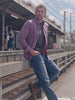 A man standing outdoors near train tracks wearing a purple and red striped button-down shirt over a black t-shirt and blue jeans